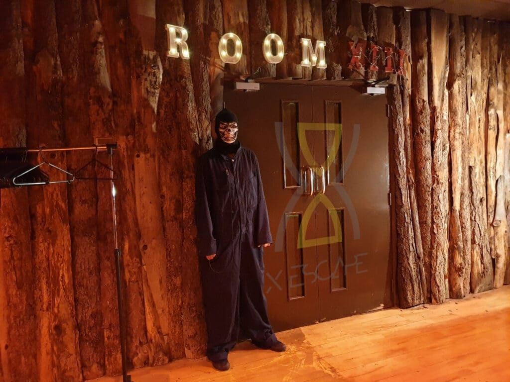 فردی با لباس و نقاب مشکی در کنار دری با علامت «اتاق فرار ترسناک» با نورپردازی تزئینی و پوشش چوبی روستایی ایستاده است.
